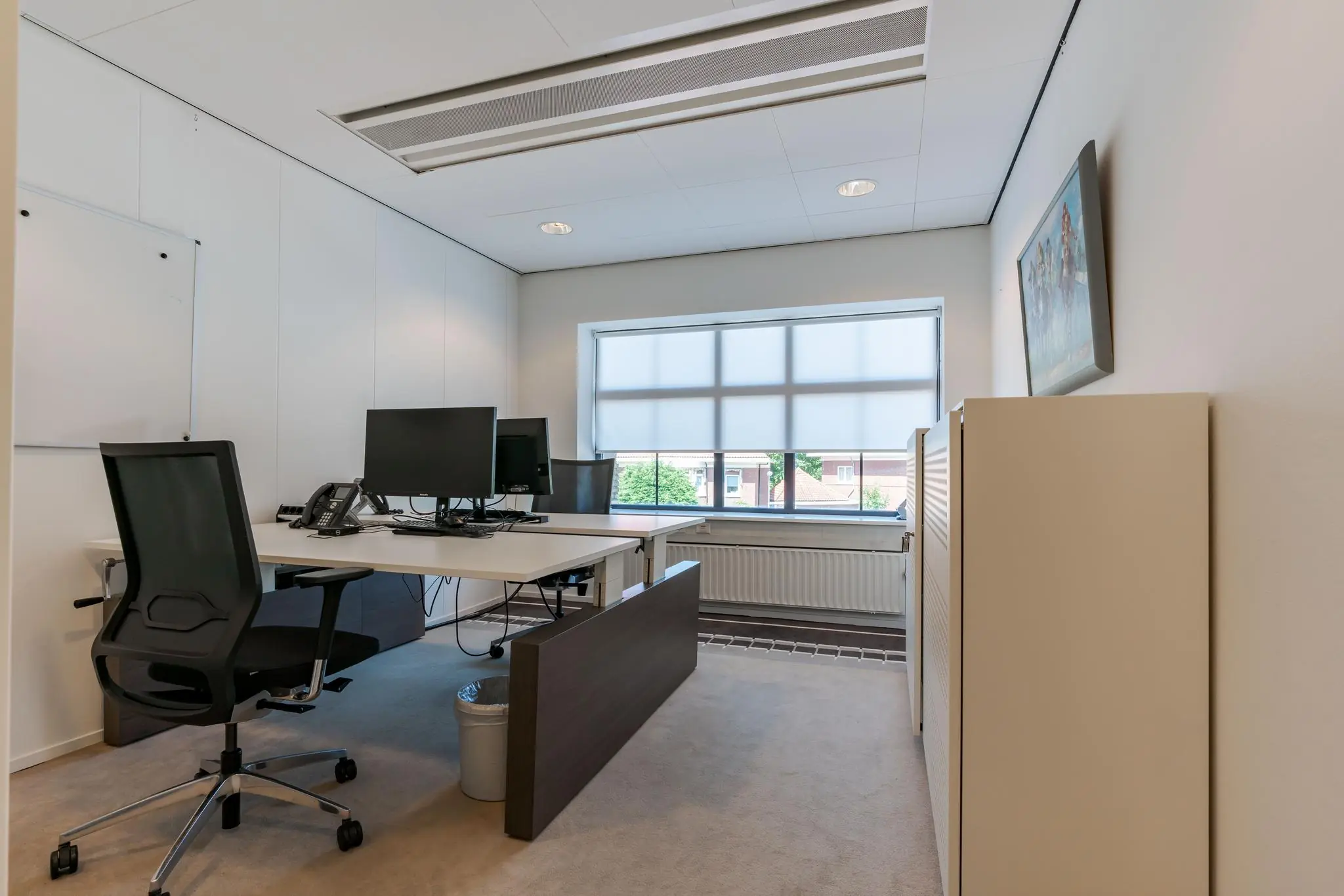 Modern ingericht kantoor aan de Van Alkemadelaan met twee bureaus, beeldschermen en een groot raam met rolgordijn.